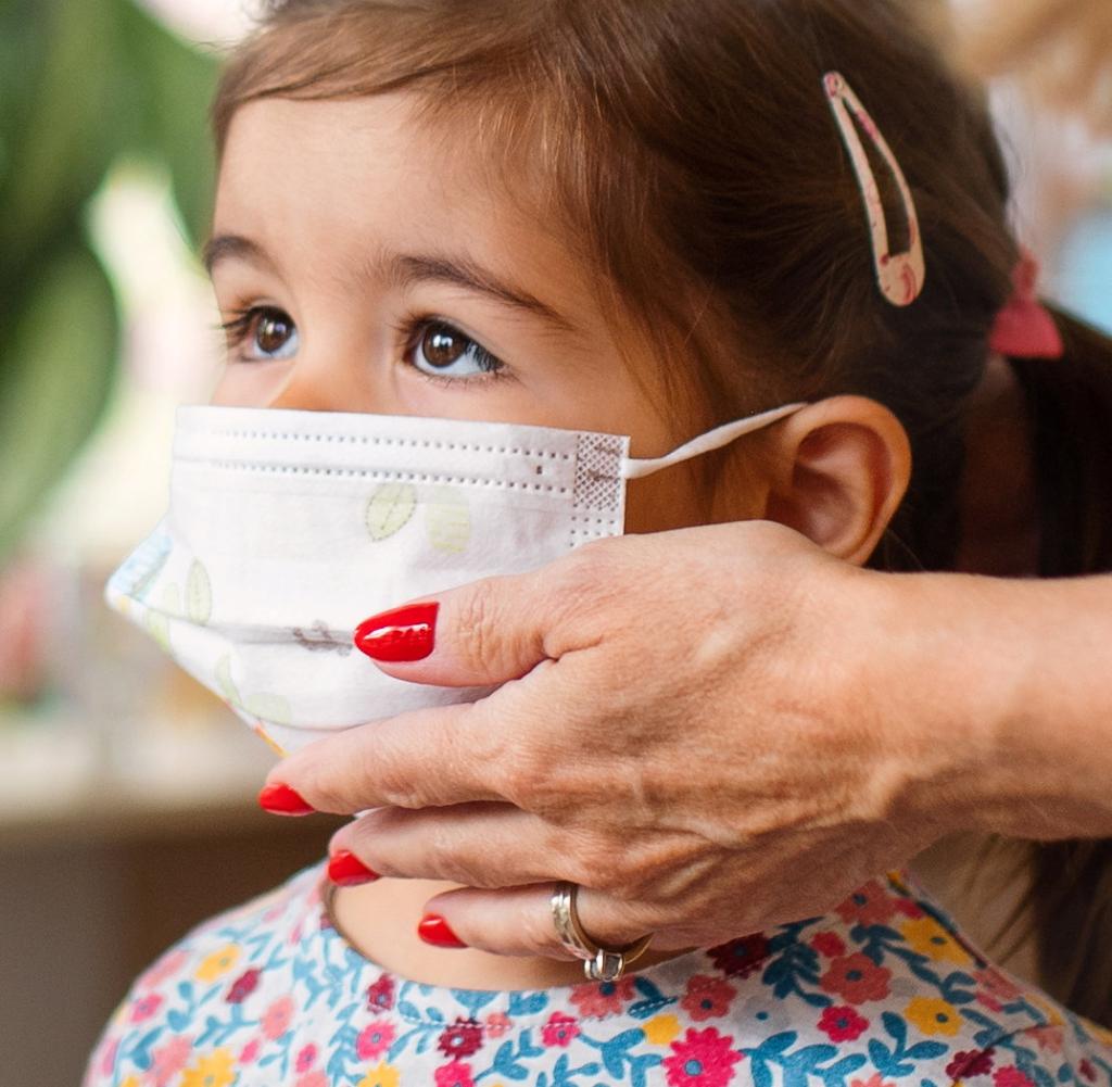 Pre-school teachers help children wear face masks indoors at nursery school, coronavirus concept.